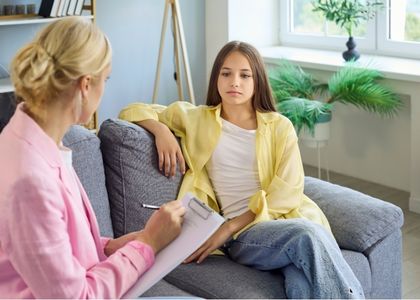 Adolescente em atendimento neuropsicológico sentada no sofá, enquanto profissional anota observações em prancheta, ambiente clínico acolhedor com estante e plantas ao fundo, fotografia realista