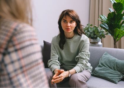 Adulto em sessão de avaliação neuropsicológica, sentado em sofá conversando com a terapeuta, ambiente acolhedor com plantas e cortinas claras ao fundo, fotografia realista