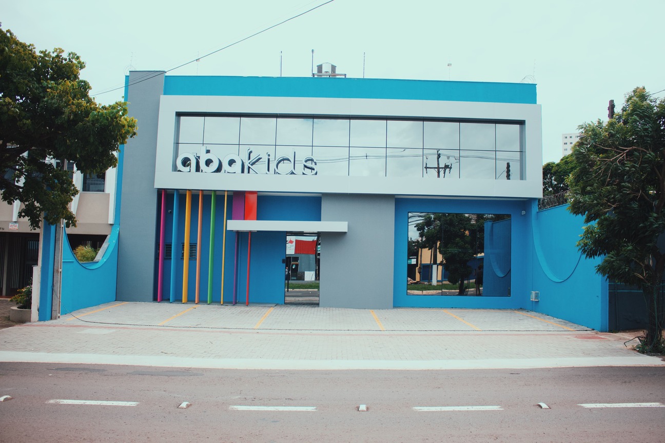 Fachada moderna da clínica ABAKIDS em Londrina, prédio azul vibrante com letreiro colorido na entrada de vidro, pilares verticais em cores variadas, estacionamento amplo à frente, fotografia arquitetônica realista em dia nublado