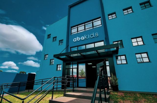 Fachada da unidade de intervenção continuada da clínica ABAKIDS, prédio azul vibrante com letreiro colorido na entrada de vidro, estacionamento amplo à frente, fotografia arquitetônica realista