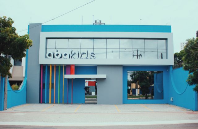 Fachada da unidade de intervenção precoce da clínica ABAKIDS, prédio azul vibrante com letreiro colorido na entrada de vidro, pilares verticais em cores variadas, estacionamento amplo à frente, fotografia arquitetônica realista